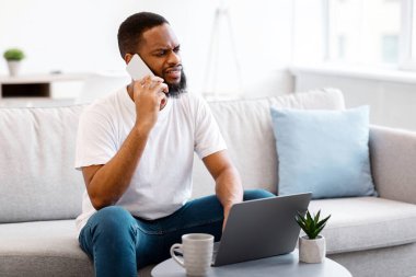 Cep telefonuyla konuşan Afrikalı adam dizüstü bilgisayarda otururken evde müşteri destek hattıyla sorun yaşıyor. Hoşnut Olmayan Müşteri ve Bilgisayar Konsepti. Seçici Odaklanma