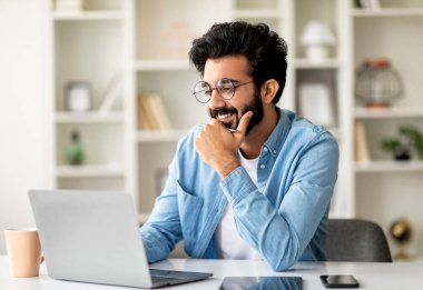 Telekomünikasyon konsepti. İçişleri Bakanlığı 'nda dizüstü bilgisayarla çalışan gülümseyen Hintli adam, masasında oturan ve bilgisayarını uzaktan kumanda için kullanan, gözlüklü yakışıklı Doğulu adam.