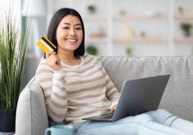 Çevrimiçi bankacılık kavramı. Dizüstü bilgisayarı ve kredi kartı olan mutlu bir Asyalı kadının portresi oturma odasında oturmuş, gülümseyen Koreli bir kadının evden ödemeleri yaparken eğlenmesi, bedava alan...