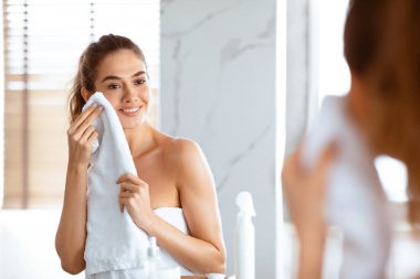 Evdeki modern banyoda aynanın yanında durduktan sonra yumuşak beyaz havluyla kuruyan genç bir kadın. Yüz temizliği ve cilt bakımı, Sabah Güzelliği rutini. Seçici Odaklanma