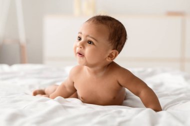 Çocuk bakımı konsepti. Yatak odasındaki beyaz battaniyenin üzerinde sürünen küçük siyah bebek portresi. Çıplak, meraklı afro çocuk karnının üstünde yatıp başka tarafa bakıyor. Boş kopyalama alanı, bulanık arkaplan