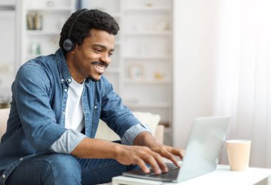 Kot ceketli, gülümseyen siyah bir erkek. Yanında kahve fincanıyla birlikte beyaz bir masada dizüstü bilgisayar kullanıyor..