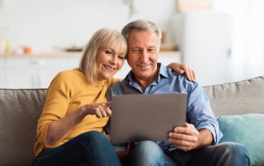 Laptop kullanan, internetten alışveriş yapan ve internette haber okuyan, evdeki koltukta oturan mutlu son sınıf çifti. Emeklilik Yaşam Tarzı, İnsanlar ve Gadget