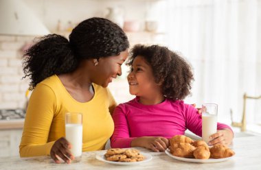 Mutfakta abur cubur yerken mutlu siyah anne ve kızı, neşeli Afrikalı Amerikalı annesi ve küçük çocuğu masada oturmuş kurabiye yiyor ve süt içiyor, bedava uzay boşluğu.