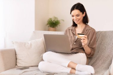 Çevrimiçi Mağaza. Mutlu kız internetten alışveriş yapıyor. Laptop ve kredi kartı kullanıyor. Kanepede oturuyor.