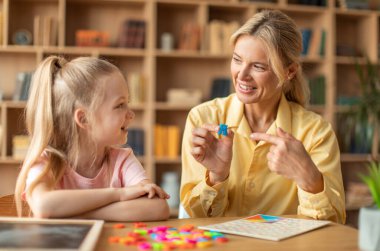 Happy woman speech therapist teaching little girl with pronounciation deffects to say sound R during personal training at classroom
