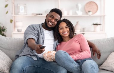 Patlamış mısırlı romantik siyah çift evde televizyon izliyor, kameraya sarılıyor ve gülümsüyor.