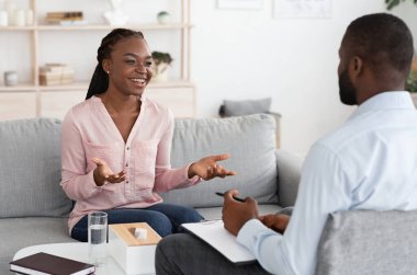 Mutlu Afrikalı kadın özel terapi seansında psikologla konuşuyor, ilerlemesini paylaşıyor, kanepede oturuyor ve gülümsüyor.