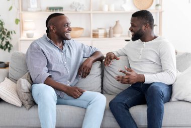 Yetişkin oğlu ve siyah babası evdeki koltukta dinlenirken konuşuyorlar, birbirlerine gülümsüyorlar, boş alan yaratıyorlar.