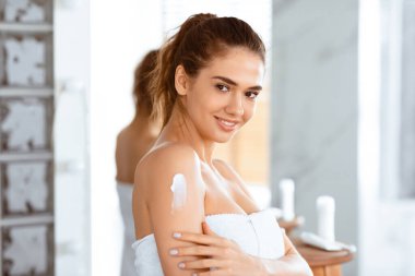 Vücut Cilt Bakımı. Omuz nemlendirici krem süren genç bir bayan modern banyoda kameraya gülümsüyor. Şımartma ve Güzellik Konsepti. Seçici Odaklanma