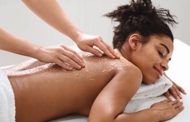 Spa terapisti siyah kadın tenini fırçalıyor, kaplıca için doğal güzellik ürünleri kullanıyor, yakın çekim.