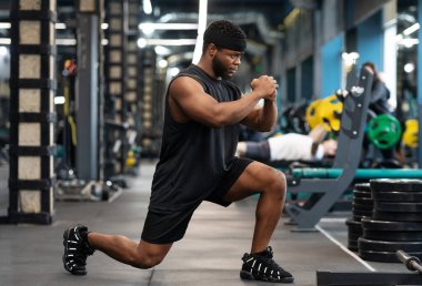 Modern spor salonunda çömelme hareketleri yapan siyahi bir sporcu, Afrika asıllı Amerikan kaslı genç vücut geliştirici, bacak kasları üzerinde çalışıyor, fotokopi alanı olan panorama, yan görüş.