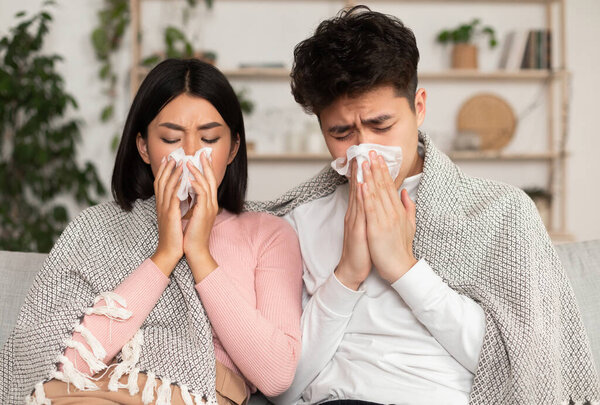 Sick Japanese Spouses Sneezing Having Cold, Blowing Nose In Paper Napkin Sitting On Couch At Home. Ill Asian Couple On Self-Isolation. Seasonal Flu, Allergy, Sinusitis And Rhinitis Illness Concept.
