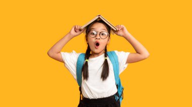 Asyalı ilkokul öğrencisi komik bir kız, sarı arka planda duran bir kitapla poz veriyor. Stüdyo Çekimi