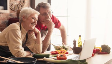 Mutfakta yemek pişirirken dizüstü bilgisayarda video tarifleri izleyen mutlu yaşlı çift, boş alanlı panorama.