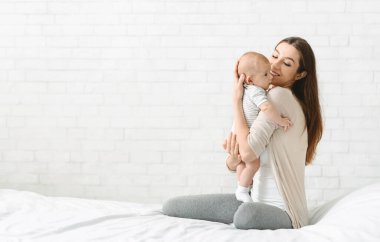 Yeni doğmuş çocuğuna sarılan mutlu genç bir kadın evde yatakta oturmuş, doğum izninin tadını çıkarıyor, boşluğu kopyalıyor.