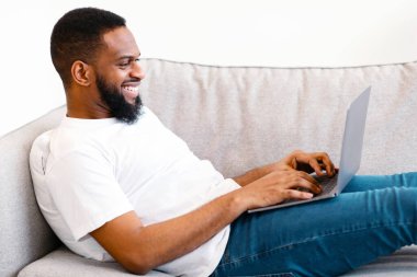 Afrikalı adam dizüstü bilgisayar kullanıyor kanepeye uzanmış internette geziniyor ve evde uzaktan çalışıyor. Kapalı alanda bilgisayar kullanan mutlu adam. Modern Teknoloji, Serbest Yaşam Tarzı Konsepti. Yan Görünüm