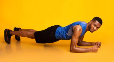 How To Do A Plank. Full-length side view of young black man in sportswear doing push ups at gym, yellow studio wall