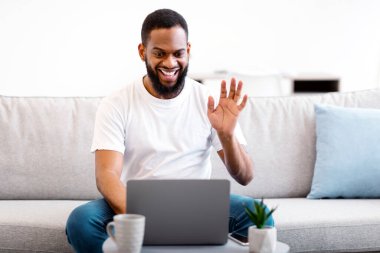 Çevrimiçi İletişim. Evdeki koltukta oturan ekranı selamlayan dizüstü bilgisayarla arayan Afrikalı adam videosu. Siyahi Adam Uzaktan Arkadaşla Video Konferansı Yapıyor.