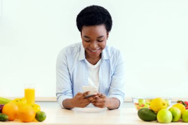 Mutlu siyahi kadın, evde modern mutfakta durup sağlıklı bir akşam yemeği hazırlarken kilo verme uygulamalı cep telefonu kullanıyor. Sağlıklı Beslenme Konsepti için Mobil Uygulama