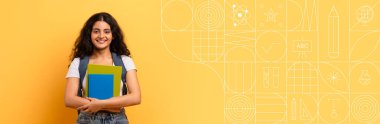 Bir öğrenci göğsüne renkli kitaplar tutarak gülümsüyor. Parlak sarı arka plan, eğitim ve öğrenim faaliyetleri için mükemmel, enerjik bir atmosfere katkıda bulunur..