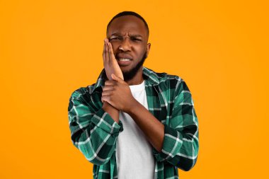 Diş ağrısı konsepti. Dişleri ağrıyan Afrikalı Amerikalı adam, sarı arka planda duran kameraya bakarken yanağına dokunuyor. Ağrıyan Diş Sağlığı Konusu Konsepti