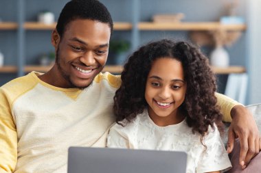 Oturma odasında kanepede otururken kızına bilgisayar kullanmayı öğreten mutlu siyah babanın portresi. Neşeli Afro-Amerikan ailesi evde dizüstü bilgisayar kullanıyor, boşluğu kopyalıyor.