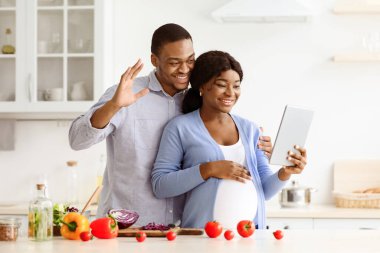 Afro-amerikan bir çiftin Kicthen 'da birlikte yemek pişirirken, dijital tablet tutarken ve Ped Screen' e el sallarken, akrabaları ya da arkadaşlarıyla görüntülü konuşma yapmasını bekliyoruz.