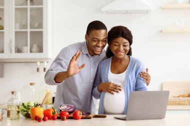Neşeli hamile bir çift, koca göbekli, yakışıklı bir erkek ve kadın. Mutfakta yemek pişirirken aileleriyle video görüşmesi yapıyorlar. Laptop ekranına el sallıyorlar.