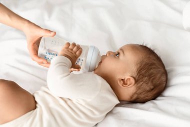 Kreş ve Çocuk Bakımı Konsepti. Süt içen küçük siyah bir çocuğun portresi, bebek bakıcısı elinde şişeyle evde yatak odasındaki beyaz battaniyenin üzerinde yatan çocuğu besliyor.