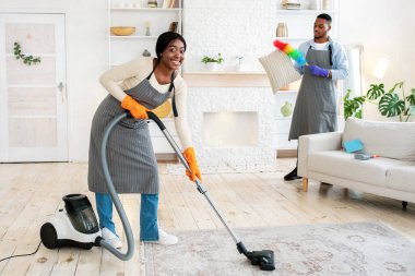 Erkek arkadaşı dairelerinde toz yastığı temizlerken yerleri süpüren mutlu siyahi kadın. Milenyum Afrikalı Amerikalı çift evi birlikte temizliyor, kopyalama alanı