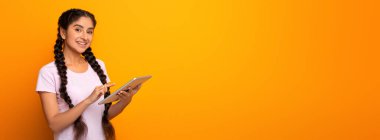 Dijital tablet tutan ve gülümseyen mutlu Hintli genç bayan portresi sarı stüdyo arka planında tek başına duruyor. Modern aletler kullanan neşeli kız öğrenci, internette çalışıyor.