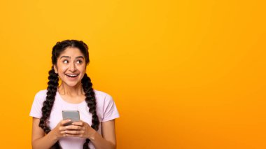 Halk ve Teknoloji Konsepti. Gülümseyen heyecanlı Hintli kadın portresi sarı turuncu stüdyo arka planında izole bir şekilde duran akıllı telefon kullanıyor ve fotokopi alanına bakıyor.