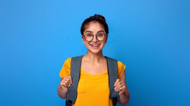 Tecritten sonra okula dön. Sırt çantası ve gözlük takan mutlu Hintli kız öğrencinin portresi mor stüdyo arka plan duvarında izole bir şekilde duran kameraya bakıyor.