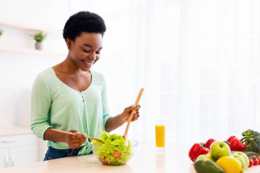 Genç Afrikalı Amerikalı kadın akşam yemeği için salata yapıyor evde yemek hazırlıyor, modern mutfakta duruyor. Sağlıklı Sebze Tarifleri ve Diyet Beslenme