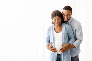Neşeli Afro-Amerikan hamile çift sarılıyor ve göbeğine bakıyor, beyaz arka planda fotokopi alanı, panorama. Zenci bir kadın ve kocasının hamileliğinin tadını çıkarmasını beklerken mutluyum.