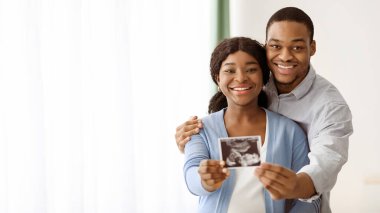 Çekici Afrikalı Amerikalı çift bebek sonogramıyla poz veriyor, ev içi, fotokopi alanı olan panorama. Mutlu siyah koca karısına sarılıyor, çocuklarının ultrason görüntülerini gösteriyor. Ebeveynlik kavramı