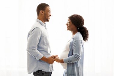 Şehvetli hamile Afro-Amerikan çift evde birlikte vakit geçirirken kucaklaşıyorlar, beyaz arka plan, fotokopi alanı. Pencerenin yanında duran ve koca göbekli kadına sarılan siyah bir çift beklerken mutlu oluyorum.