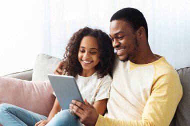 Mutlu siyah baba ve kızı oturma odasında kanepede otururken dijital tablet kullanıyor. Neşeli Afro-Amerikan aile babası ve kızı evdeki bloknotu kullanarak, boşluğu kopyalayarak