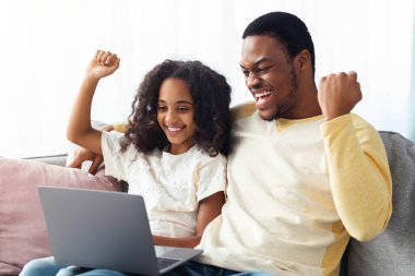 Duygusal siyah baba ve kızı oturma odasında kanepede otururken bilgisayar kullanıyor. Afro-amerikan aile kazananları evde dizüstü bilgisayar kullanıyor, video oyunları oynuyor, boşluk kopyalıyor