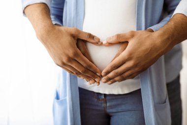 Kalbi olan siyah bir çiftin elleri hamile bir kadının karnının üzerinde, yakın plan, kopyalama alanı. Tanınmayan Afro-Amerikan ailesi bekliyor, beyaza sarılıyorlar, göbeğin üstünde el ele tutuşuyorlar.