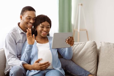 Neşeli, hamile, yakışıklı, koca göbekli, kanepede oturan ve aileleriyle veya arkadaşlarıyla video görüşmesi yapan, dijital tablet ekranına el sallayan, ev içi, uzay fotokopisi çeken bir çift.