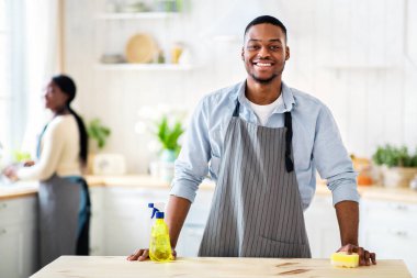 Önlüklü çekici siyahi adam portresi ve mutfakta kameraya gülümseyen lastik eldivenler, arka planda bulaşıkları yıkayan karısı, fotokopi alanı. Milenyum çifti birlikte temizlik yapıyor.