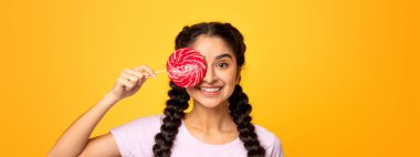 Güzel gülümseyen Hintli bir kadının portresi lolipop tutuyor, bir gözünü çubuğa şeker yapıştırıyor ve kapatıyor, parlak sarı stüdyo arka planında izole edilmiş. Eğleniyor musun, Tatlı Diş?