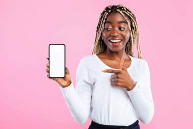 Harika Mobil Uygulama ya da Web Sitesi. Modern cep telefonuyla gülümseyen siyah bir kadının portresi beyaz ekranı işaret ediyor, cihaza bakıyor, pembe stüdyo arka planında tek başına duruyor.