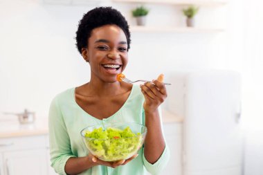 Siyah Bayan Taze Sebze Yiyiyor Sağlıklı Yiyeceklerin tadını çıkarıyor Modern Mutfakta kamera karşısında gülümsüyor. Beslenme ve kilo verme konsepti. Akşam Yemeği Yiyen Kadın İçerde Zayıflıyor