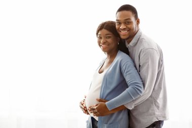 Gülümseyen siyah adam hamile, neşeli karısına beyaz arka planda sarılıyor, sahtesiyle panorama yapıyor. Mutlu Afro-Amerikan çifti evde rahat bir şekilde poz vermeyi bekliyor.