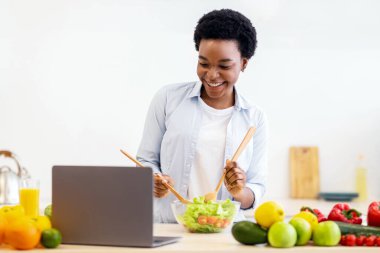 Siyahi kadın yemek pişiriyor ve dizüstü bilgisayar kullanıyor akşam yemeği için taze sebze salatası hazırlıyor ve evdeki modern mutfakta yemek tarifleri tarıyor. Sağlıklı Beslenme ve Çevrimiçi Yemek Kursları. Seçici Odaklanma