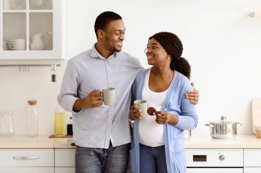 Siyah bir ailenin mutfakta kahve içmesini, sarılmalarını ve birbirlerine bakmalarını beklemekten mutluyum. Neşeli Afro-Amerikan hamile kadın ve kocası birlikte bitki çayı içiyorlar.