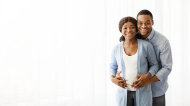 Mutlu siyah çift bebek bekliyor, beyaz arka planda poz veriyorlar, kameraya sarılıyorlar ve gülümsüyorlar, fotokopi alanı olan bir panorama. Pozitif Afro-Amerikan hamile ailesi evde sarılıyor.